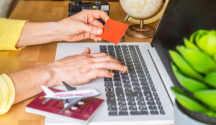 Woman is using laptop and credit card to book a flight, with miniature airplane and passport placed on the laptop, suggesting an upcoming trip